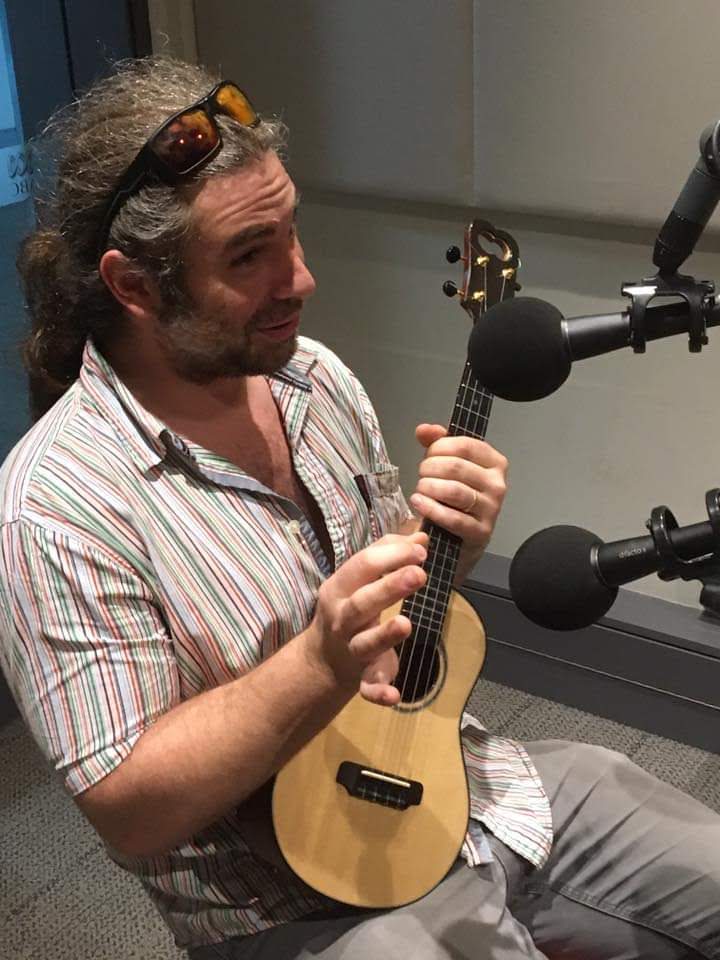 Russ speaking into microphones in a radio studio while holding a ukulele